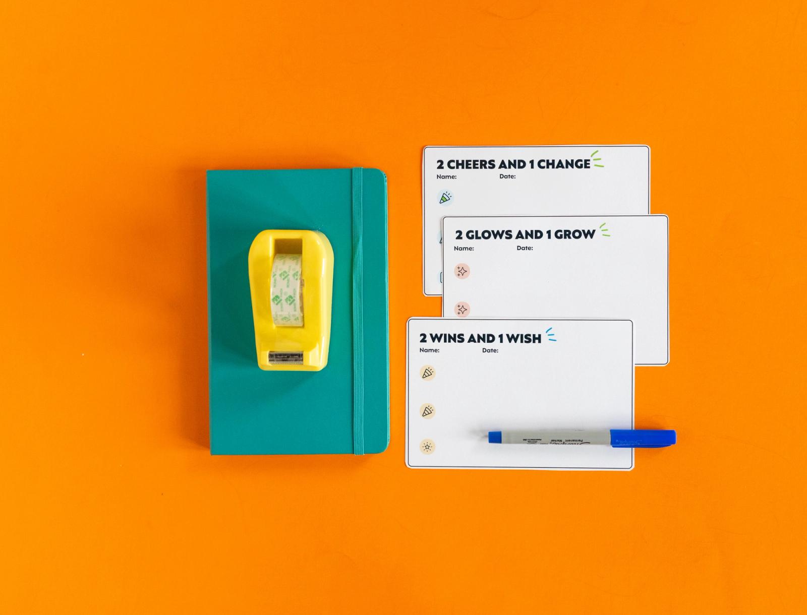 Three blank notecards and a blue pen lay on an orange background. Beside them is a teal notebook and yellow tape dispenser.