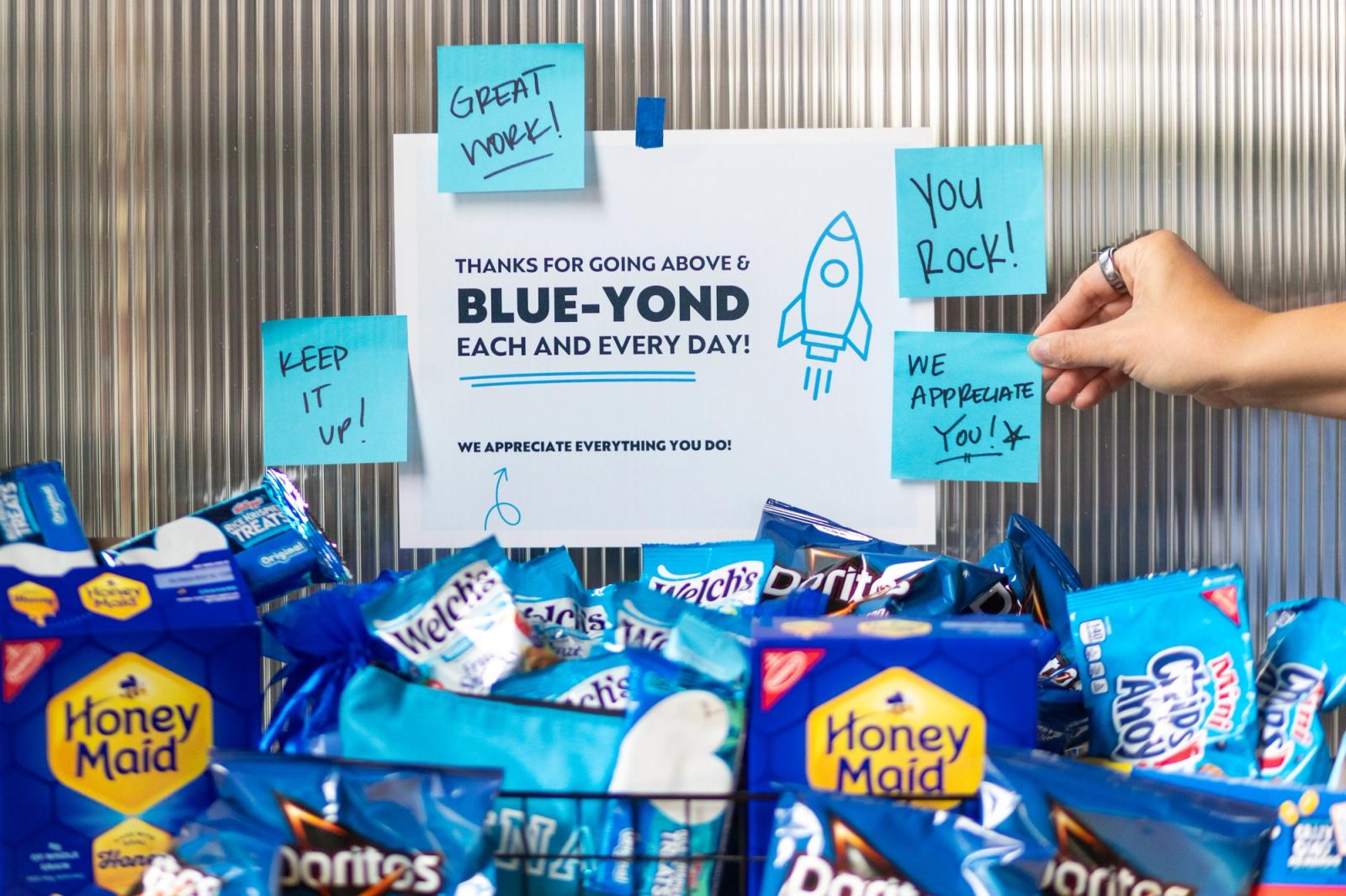 An array of snacks in blue bags sits in front of a wall with a poster and blue sticky-notes.