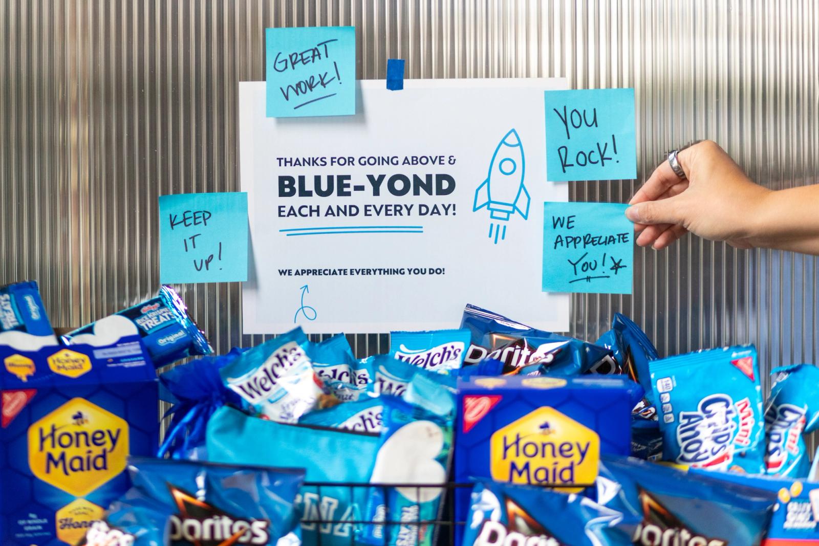An array of snacks in blue bags sits in front of a wall with a poster and blue sticky-notes.