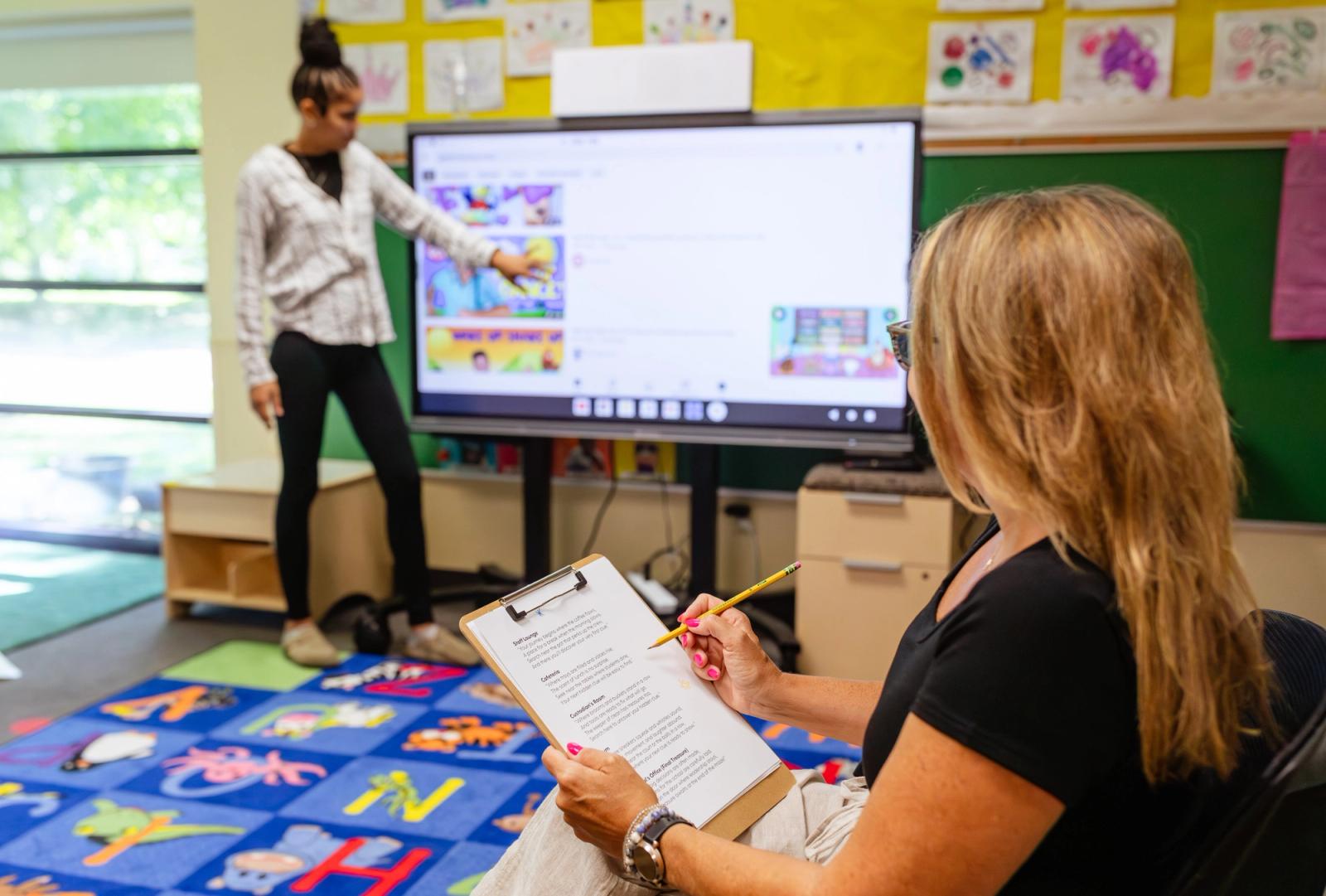 A woman stands in a classroom in front of a large television screen pointing at it. A woman sits in front of her writing on a piece of paper on a clipboard.