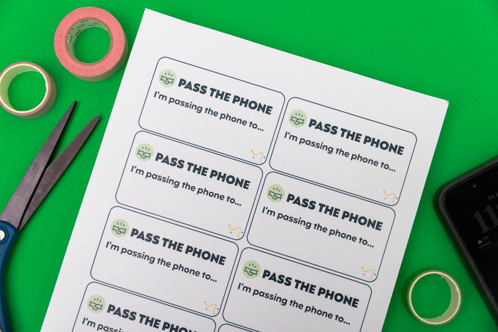 A piece of paper that says "pass the phone" scissors, and rolls of tape lay on a green background.