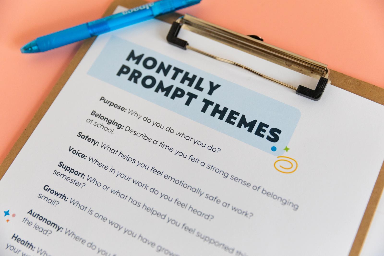 Closeup of paper that says "monthly prompt themes" on it and a blue pen on a clipboard.