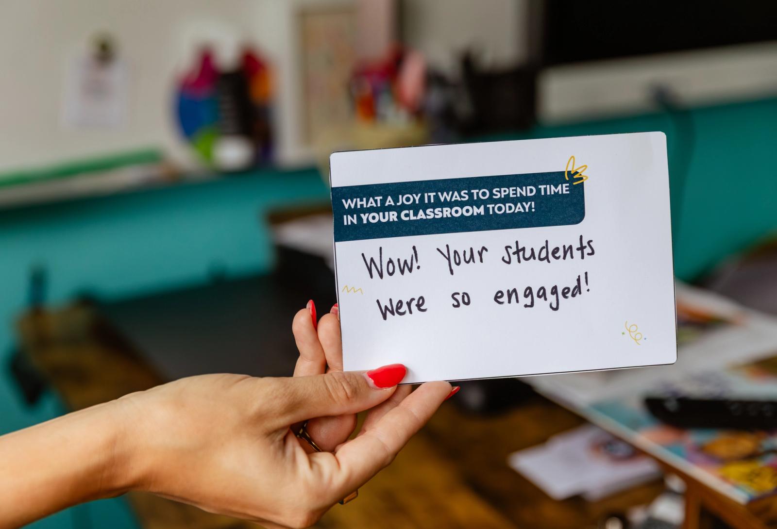A hand holds a notecard with a handwritten note on it. The background is a classroom desk and whiteboard.