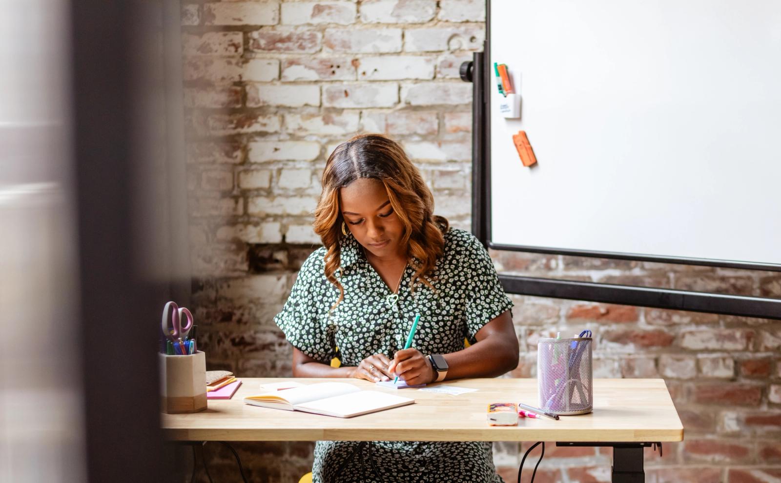 A woman sits at a desk writing on a piece of paper. A whiteboard and brick wall are in the background.