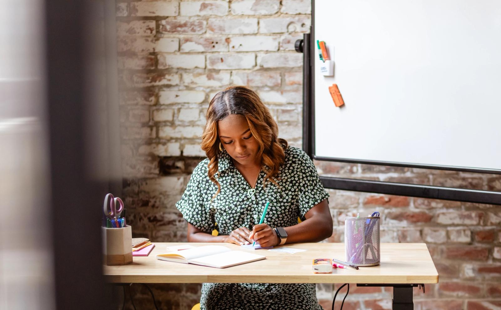 A woman sits at a desk writing on a piece of paper. A whiteboard and brick wall are in the background.