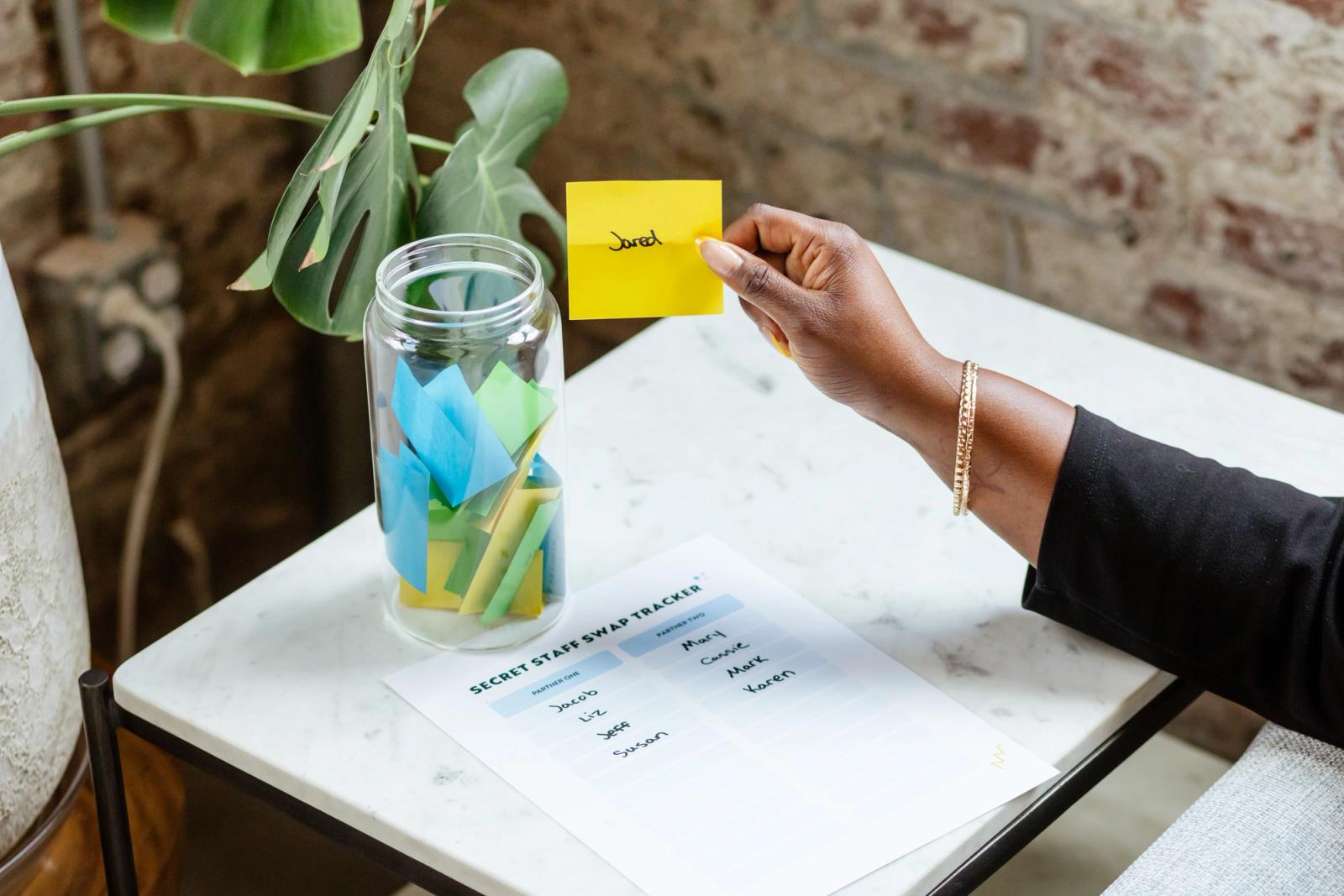 A glass jar with pieces of paper in it sits on a table next to a paper sign up sheet. A hand reached in and grabs a piece of paper with a name on it.