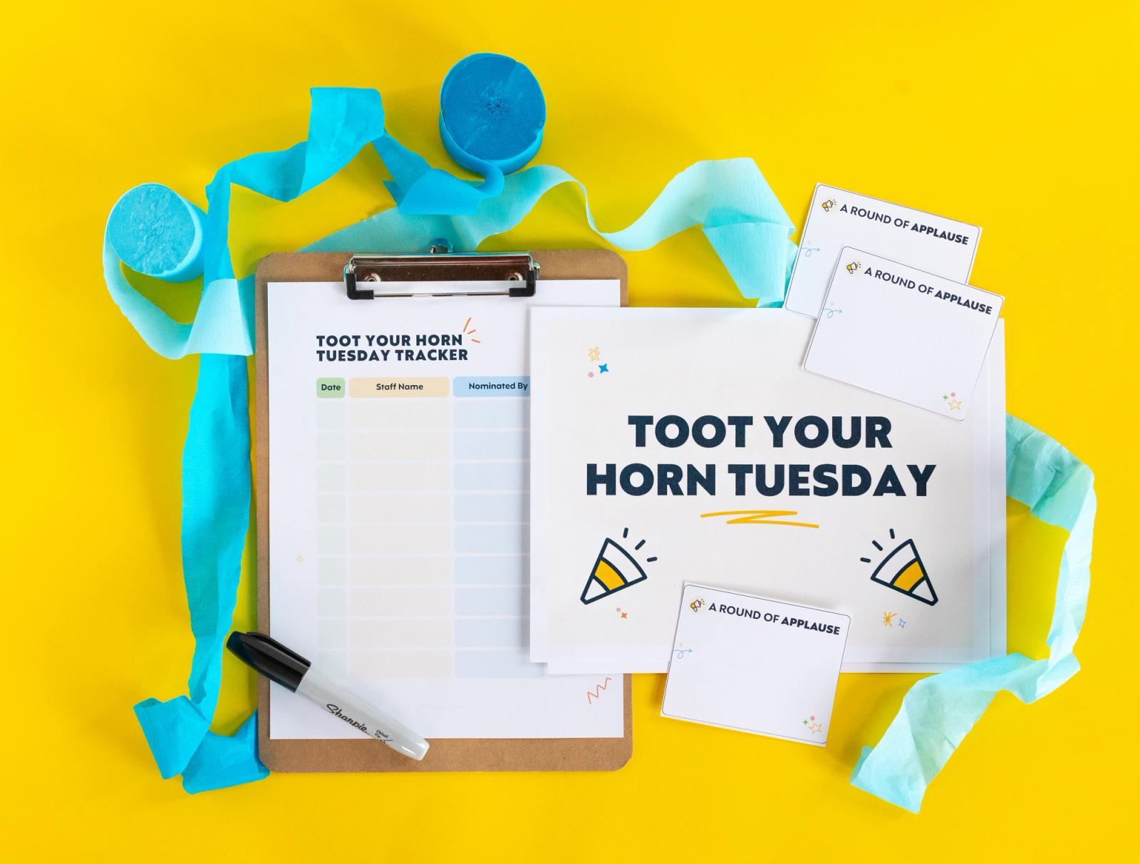 A sheet of paper that says "toot your horn tuesday" on it, clipboard, blue streamers, and maker lay on a yellow background.