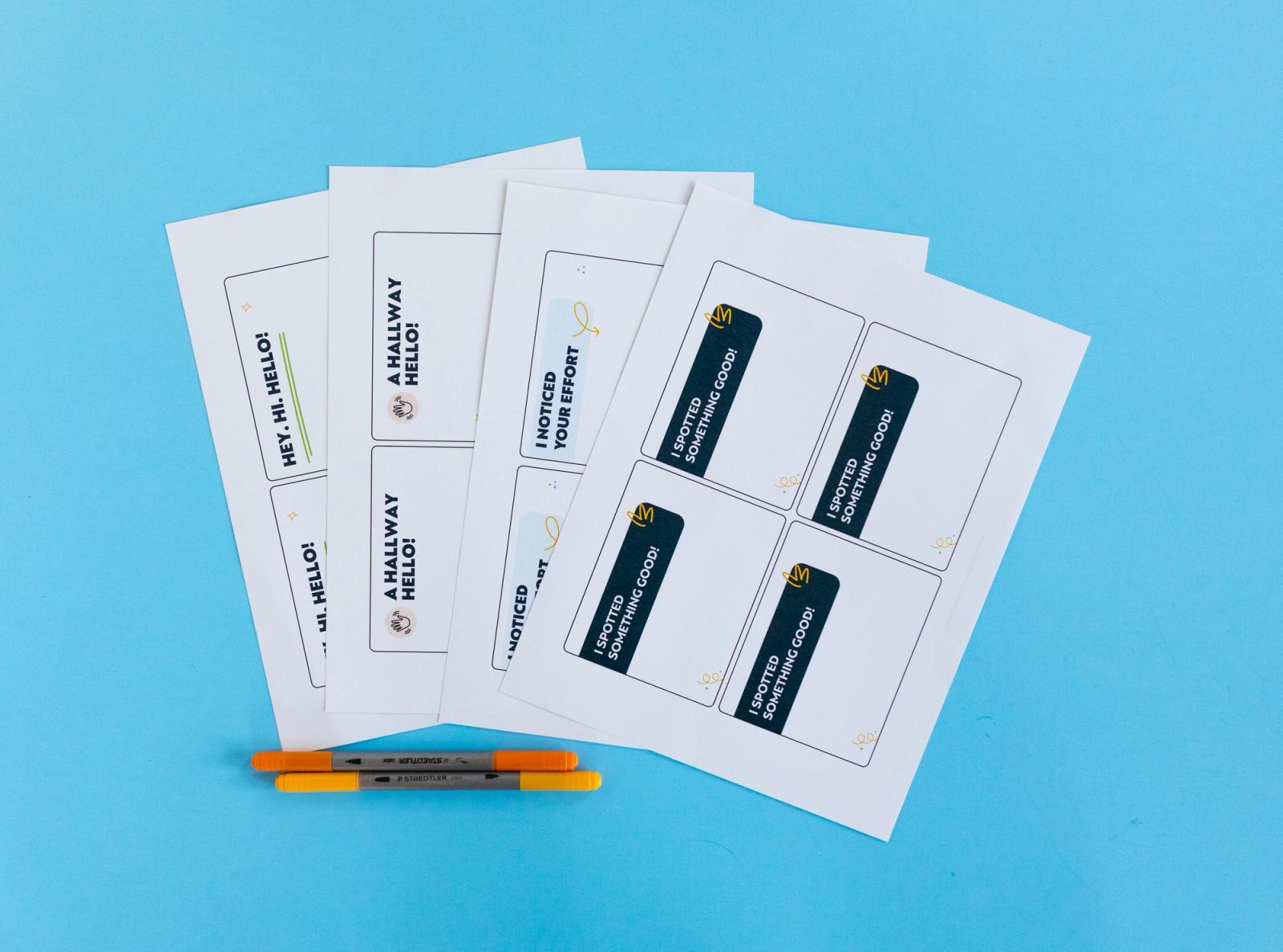 Four sheets of paper with four notecards on them lay on a bright blue background. Orange pens lay beside them.
