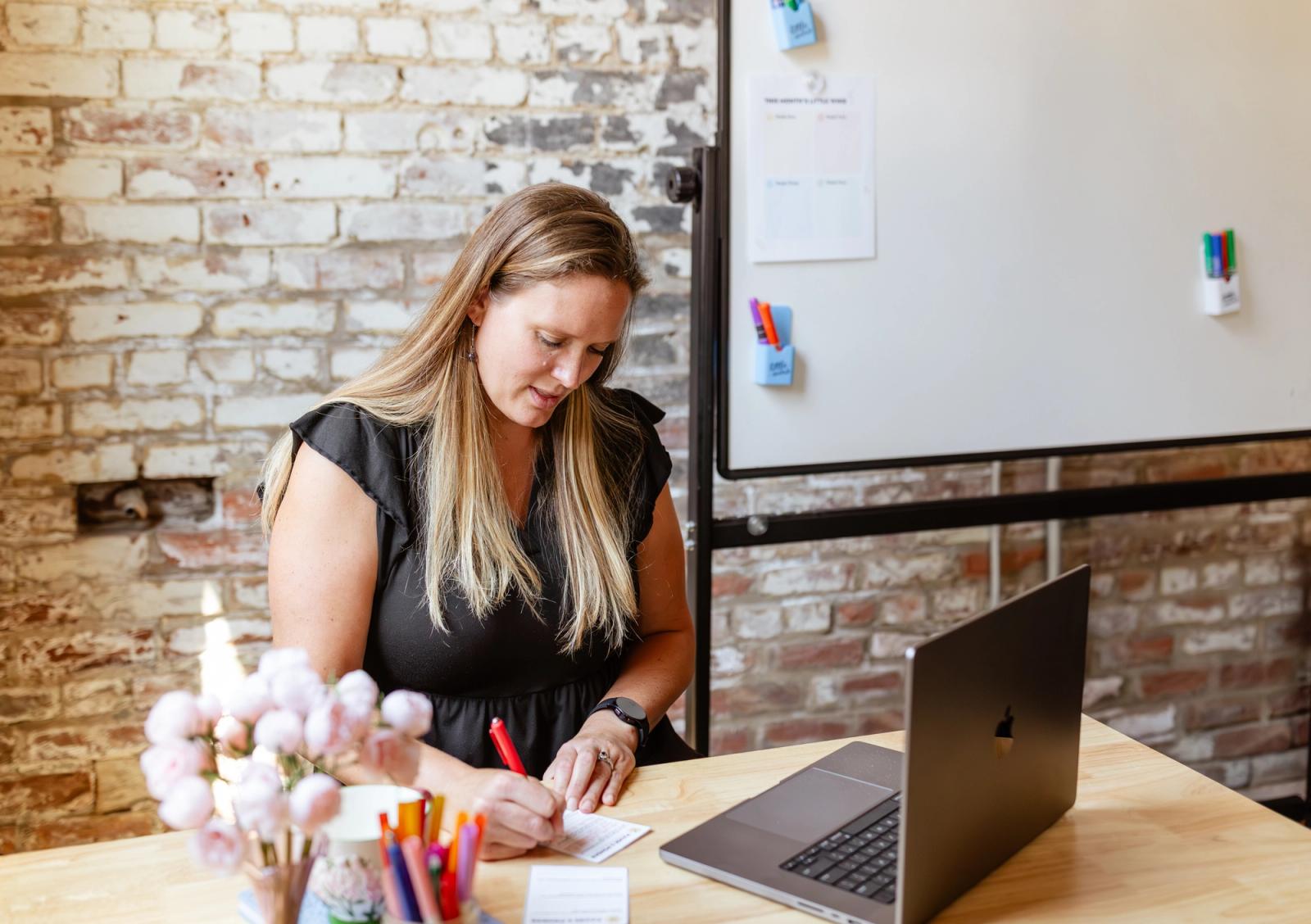 A woman sits at a desk with an open laptop on it, writing on a piece of paper. A brick wall and whiteboard are in the background.