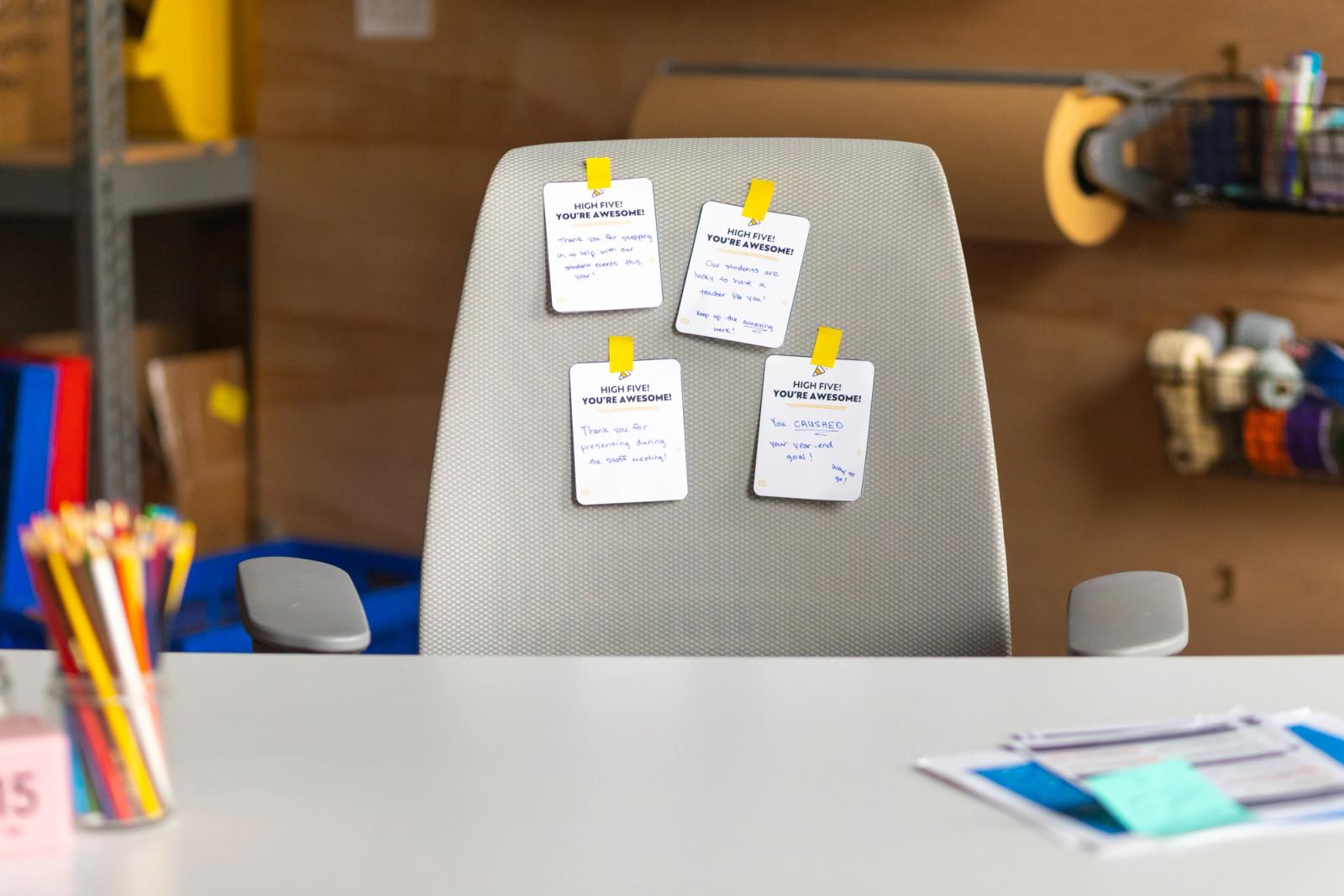 Four notecards taped to a desk chair. The chair is in front of a white desk with papers and jar of pencils.
