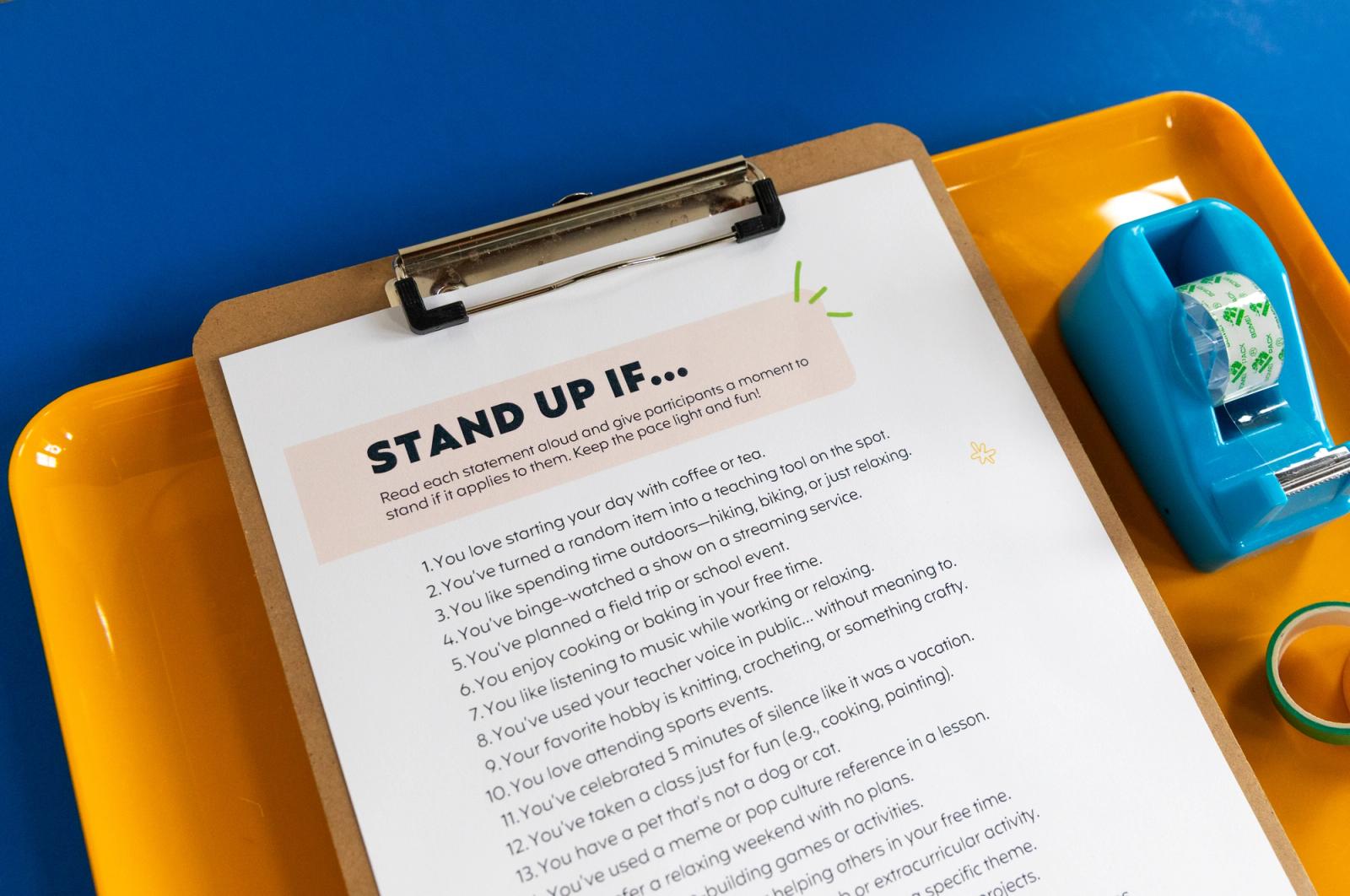Closeup of a piece of paper that says "stand up if"on a clipboard laying in an orange tray with a tape dispenser and roll of tape on a blue background.