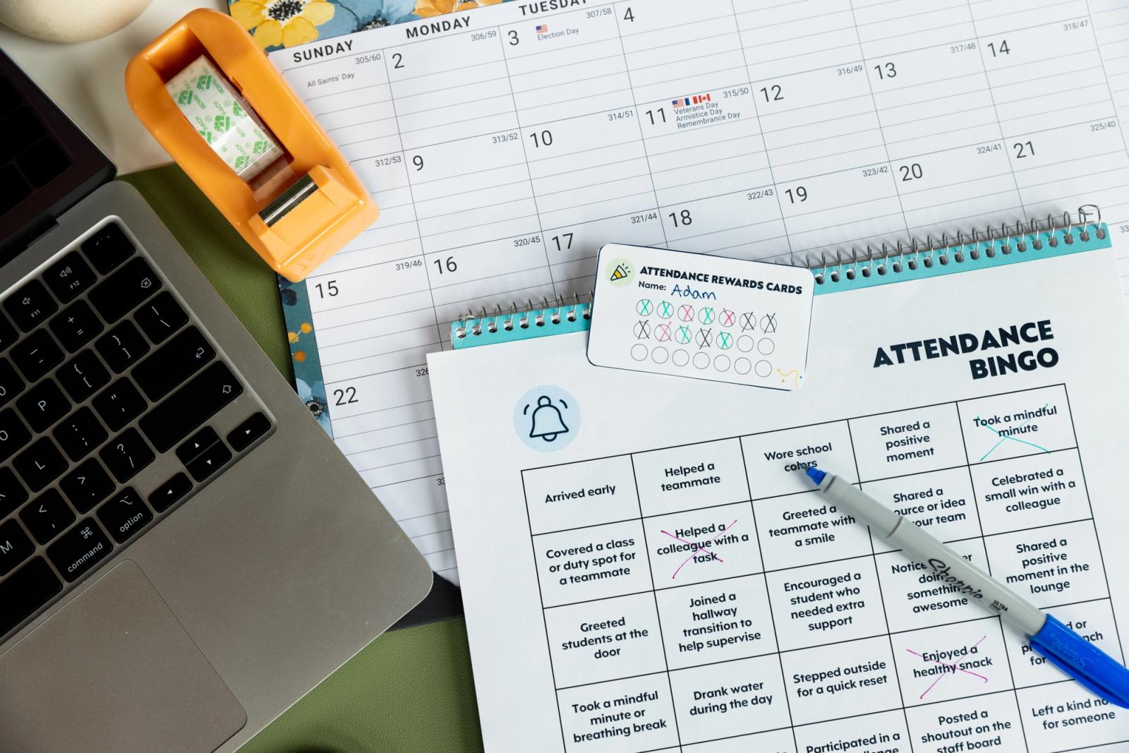 A piece of paper that says "attendance bingo" lays on a desk with a calendar and open laptop.