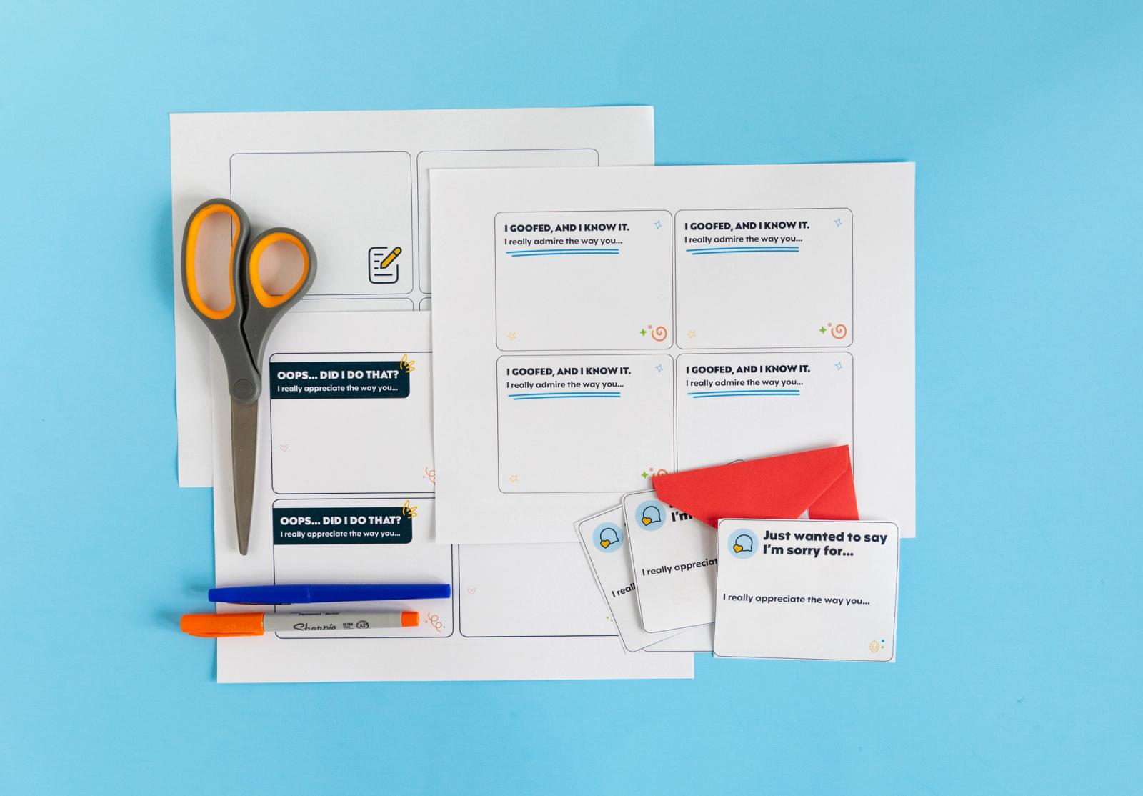 Sheets of paper, notecards, scissors, and markers lay on a light blue background.