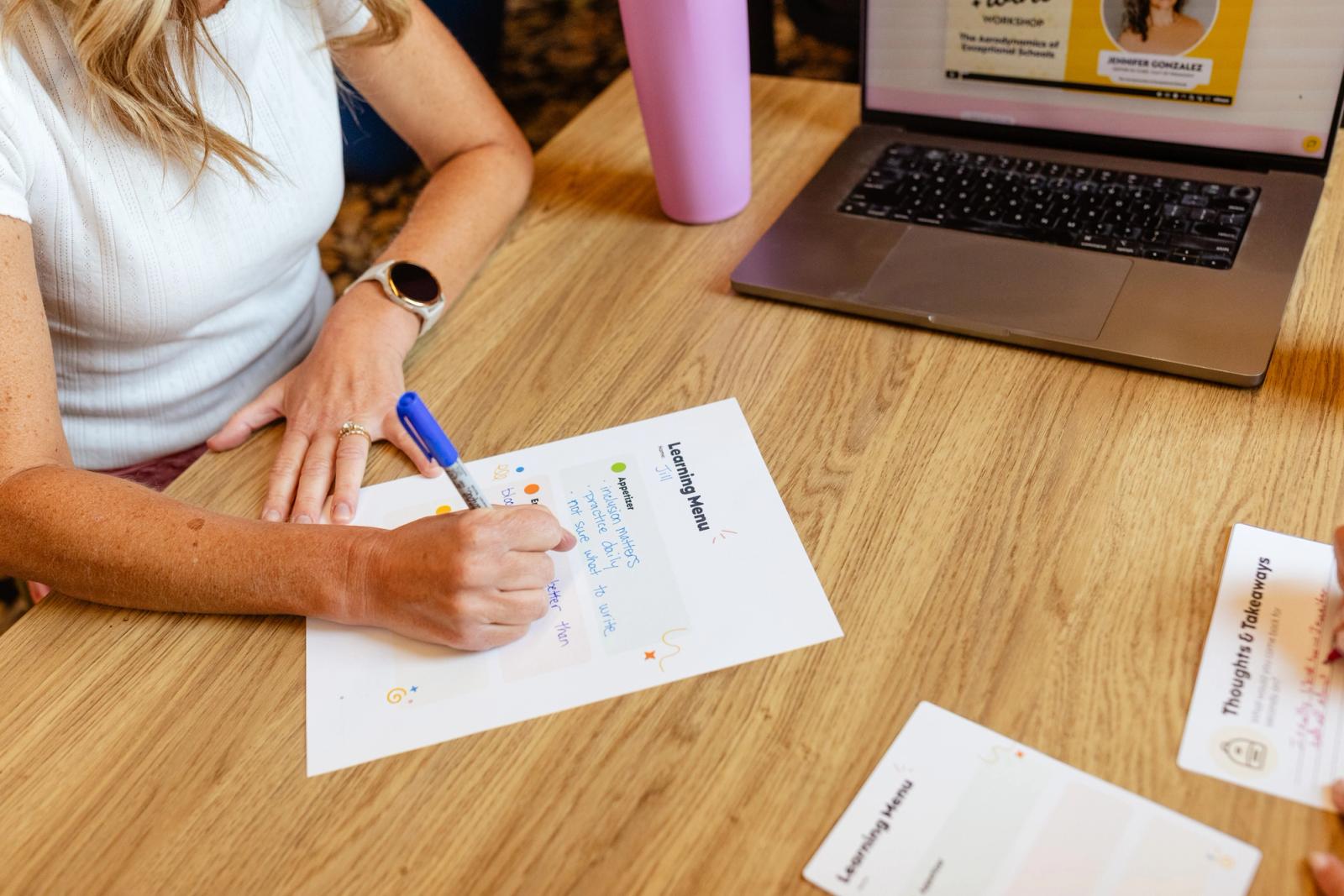 A woman sits at a table with an open laptop. She writes on a piece of paper with a blue pen.