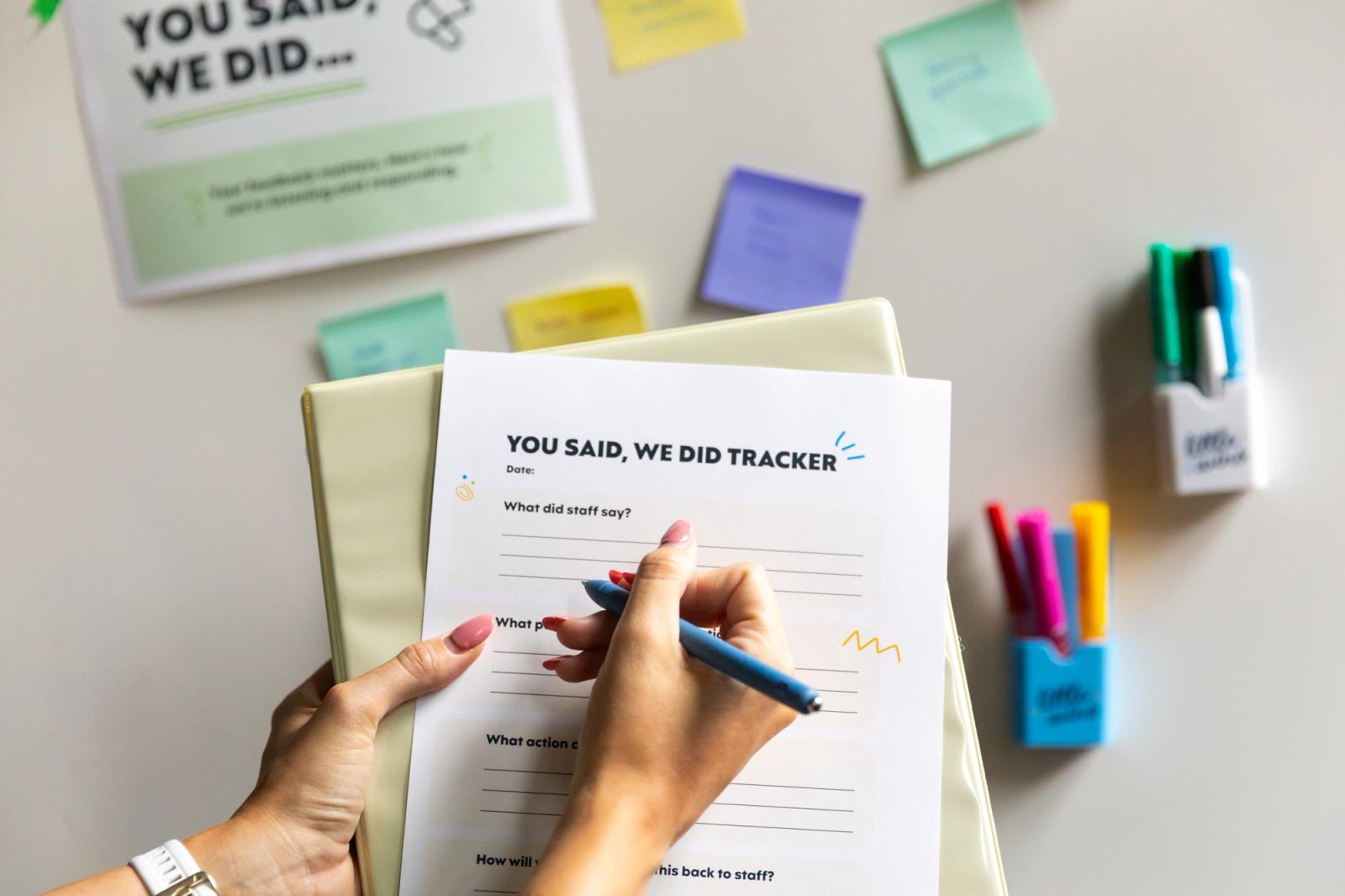 A hand writes on a piece of paper that says "you said, we did tracker." In the background is a whiteboard with colorful sticky notes and dry erase markers stuck to it.