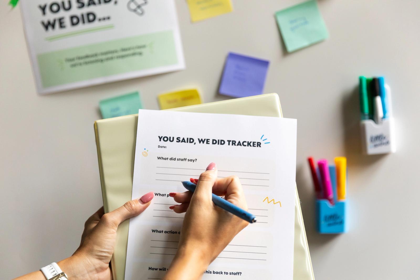 A hand writes on a piece of paper that says "you said, we did tracker." In the background is a whiteboard with colorful sticky notes and dry erase markers stuck to it.