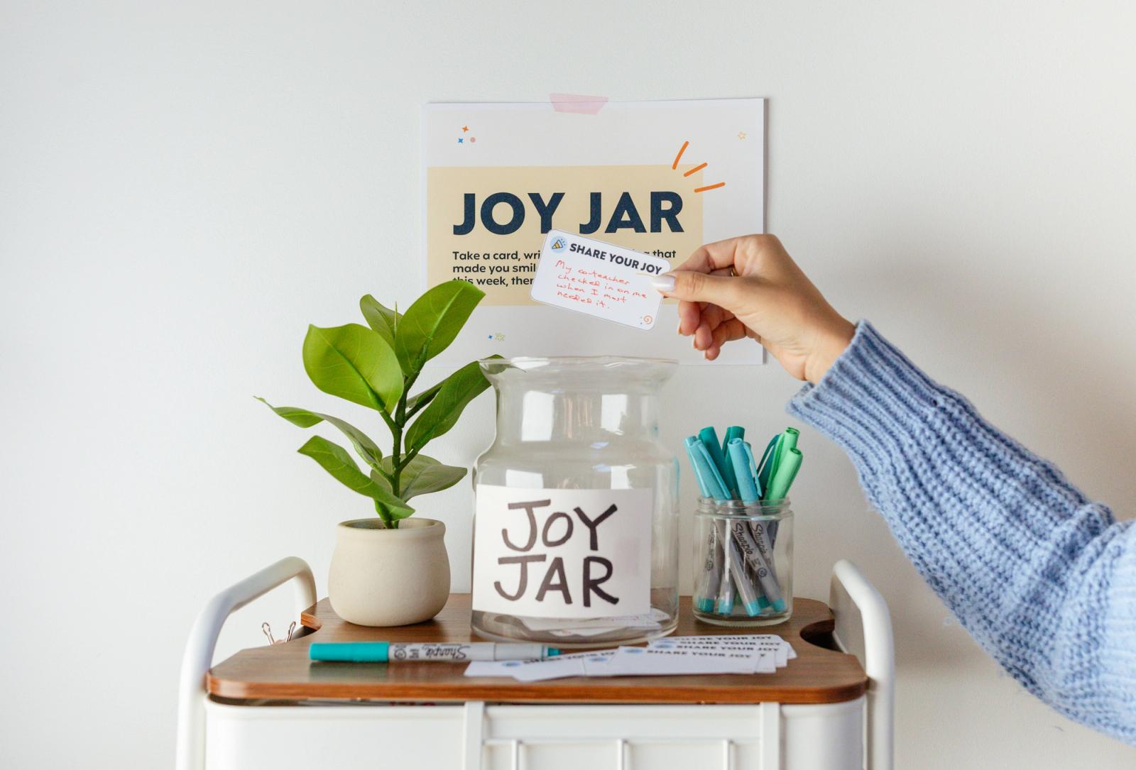 A small cart with a glass jar, plant, pens and notes sits in front of a white wall. A poster above the card says "joy jar." A hand reaches in to drop a note in the jar.
