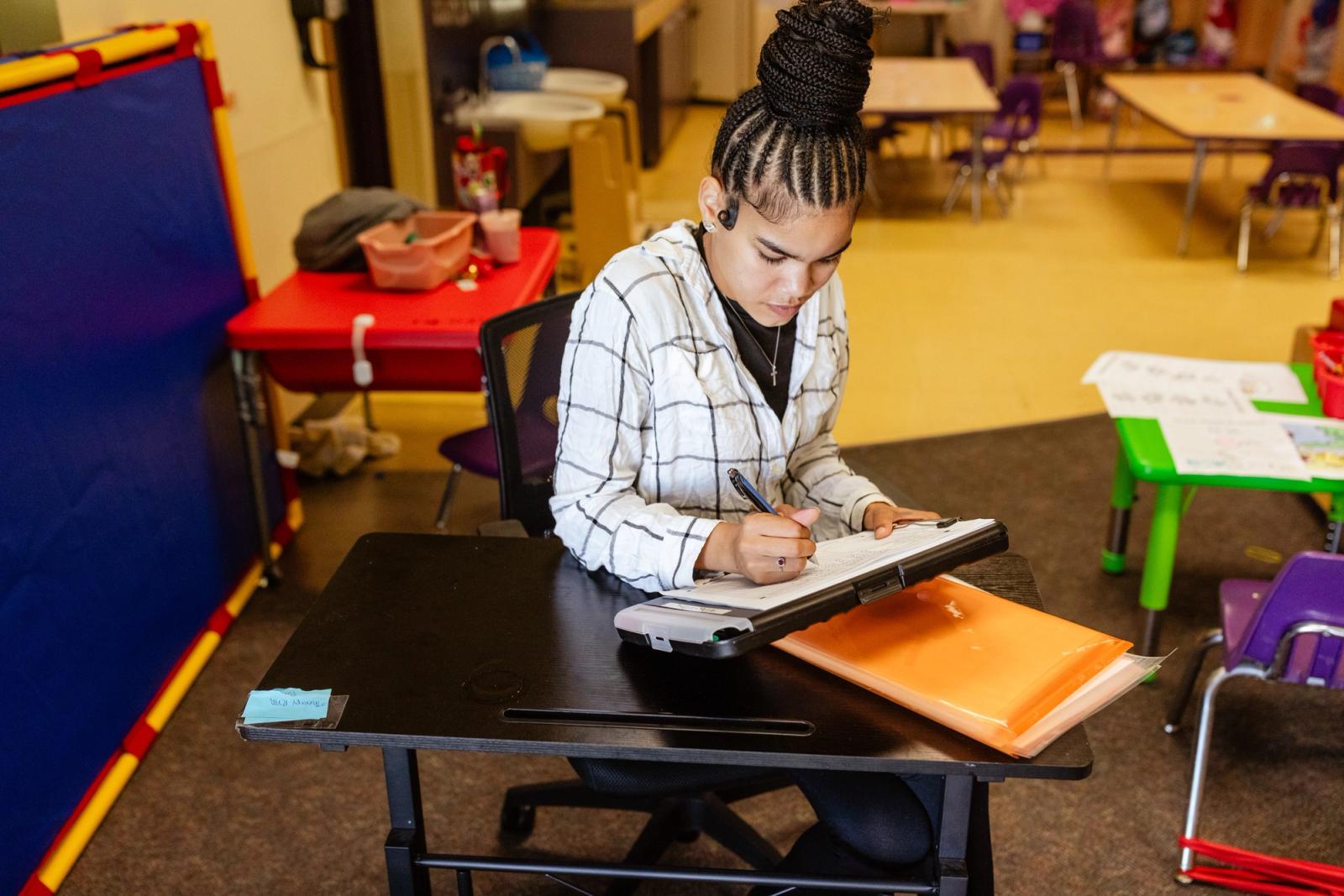 A woman sits at a small table in a classroom. She is writing on a piece of paper in front of her.