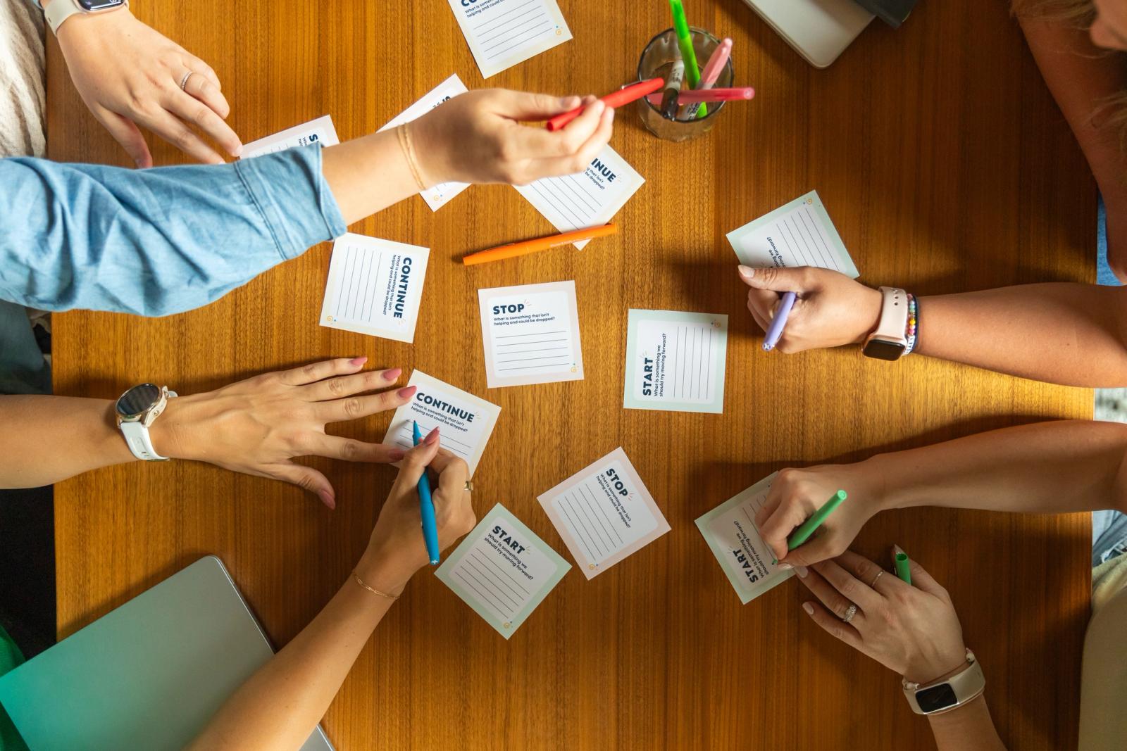 Four people sit at a brown table. They are reaching for pens and writing on pieces of paper.