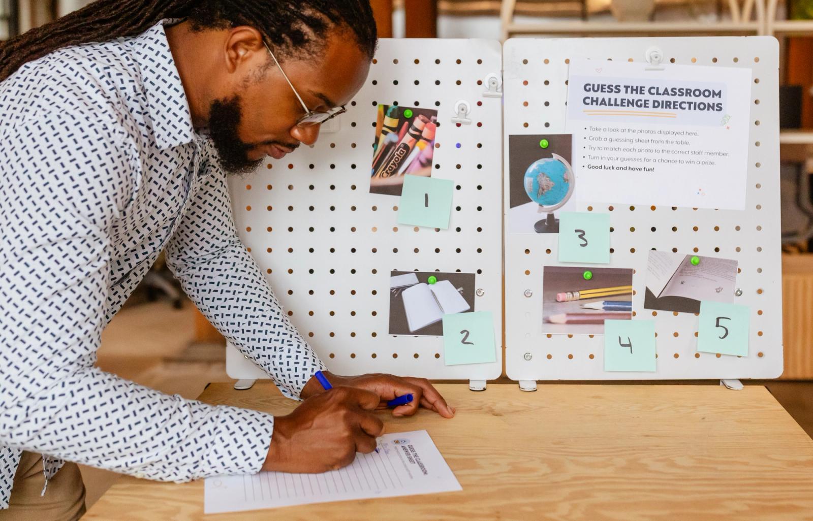 A man stands in front of a bulletin board with photos on it. He writes on a piece of paper.