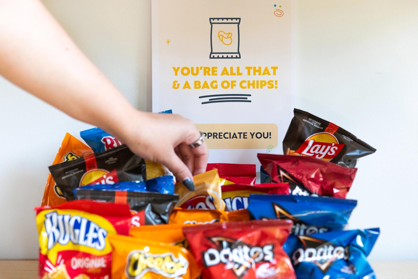 A stack of small bags of chips sits on a table with a sign posted on the wall behind them. A hand reaches in to grab a bag.