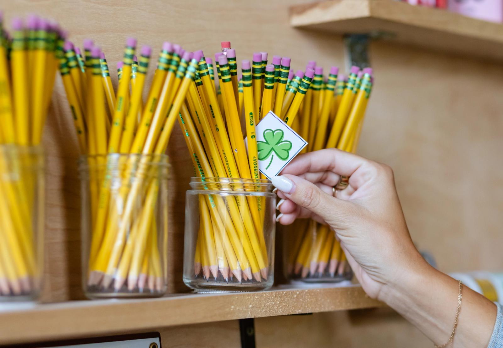 A hand places a small piece of paper with a green shamrock on it in a glass jar of pencils.