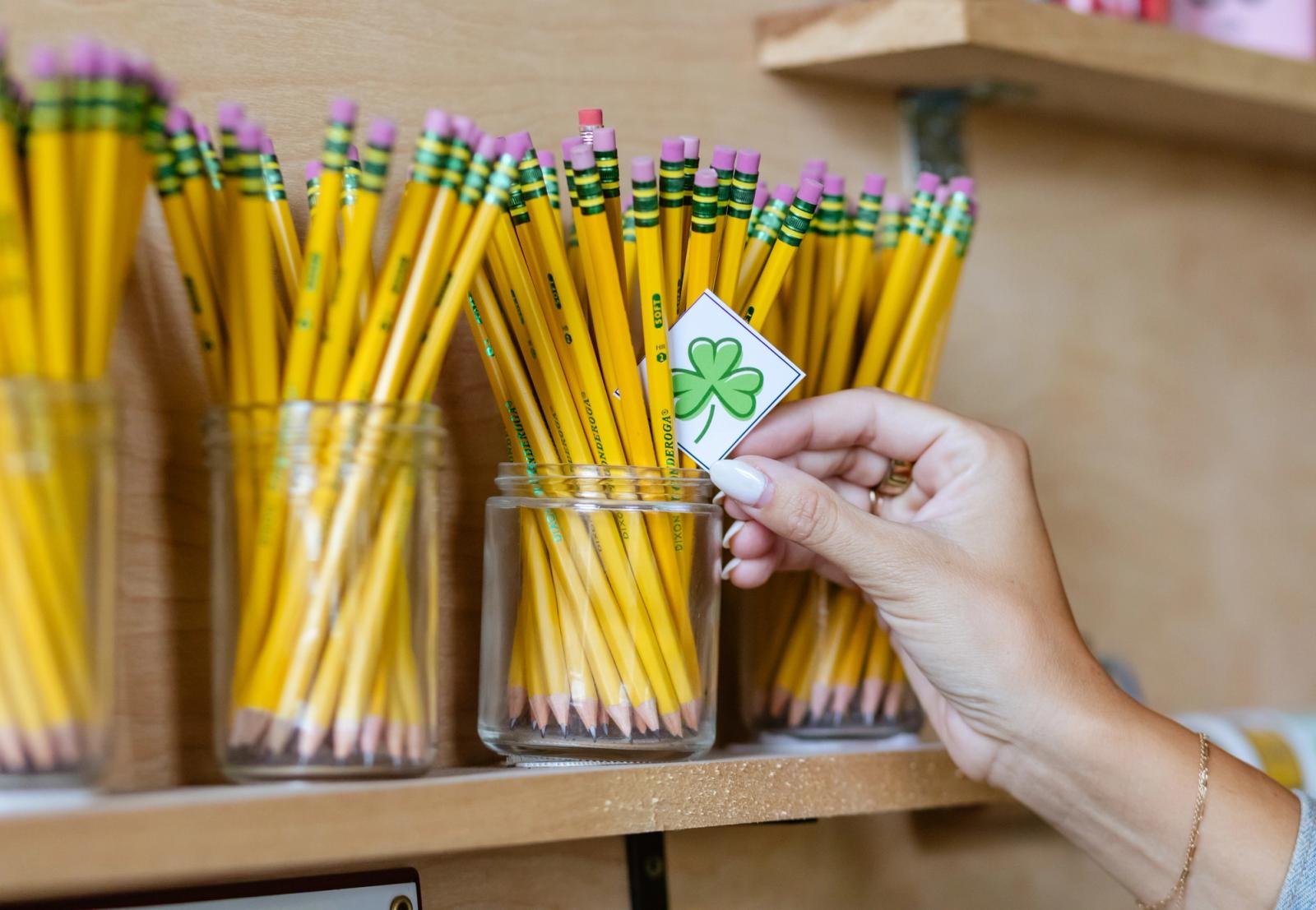 A hand places a small piece of paper with a green shamrock on it in a glass jar of pencils.