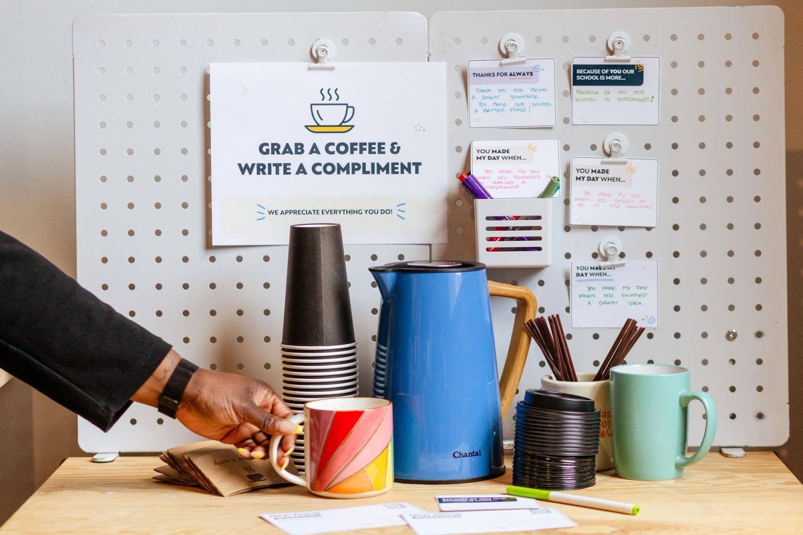 Coffee mugs, to-go cups, a kettle, and coffee supplies sit on a wooden table in front of a peg board. A poster and handwritten notes are taped to the peg board. A hand reaches in to grab a coffee mug.