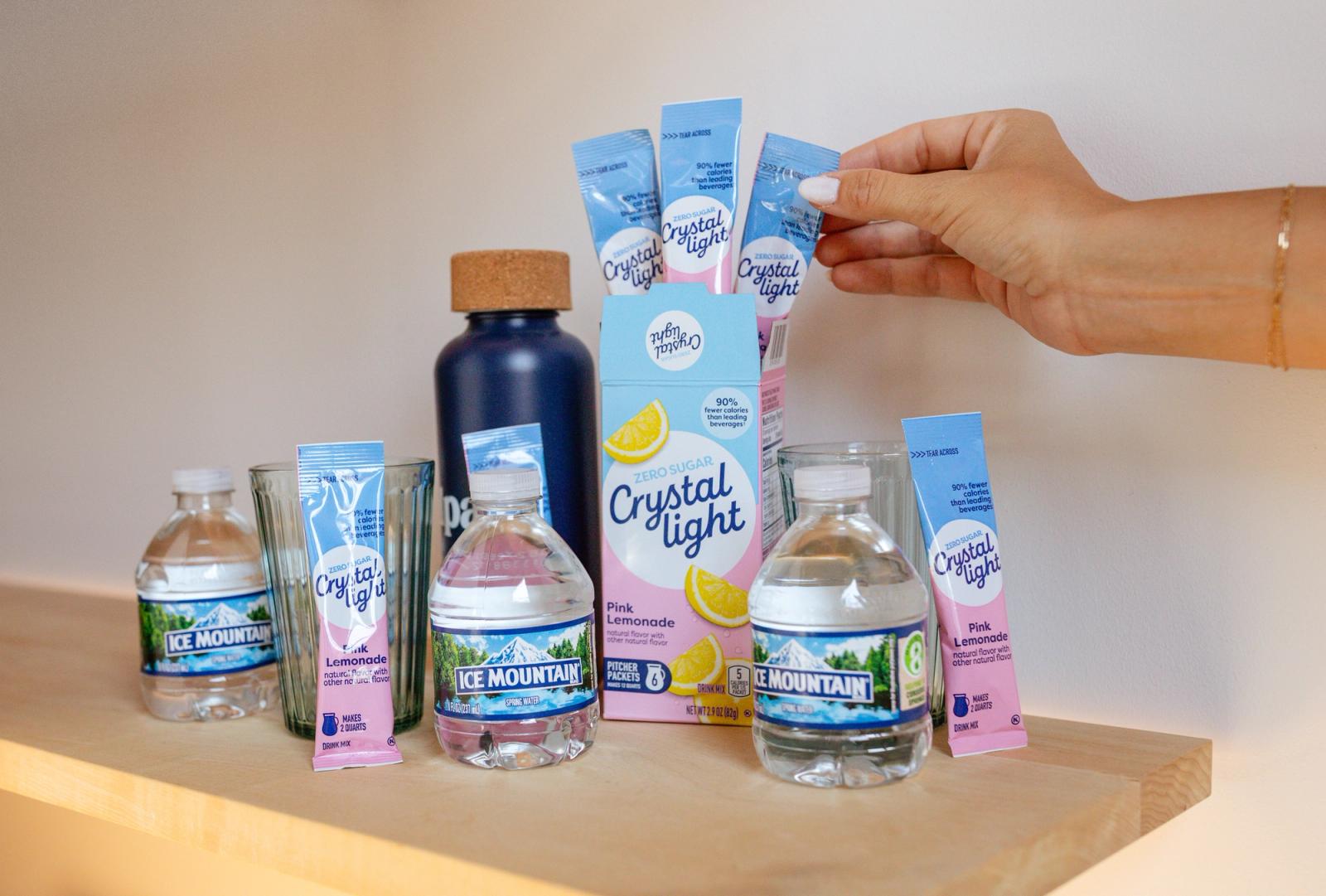Mini bottles of water, drink packets, and a water bottle sits on a wood shelf. A hand reaches in from the right to grab a drink packet.