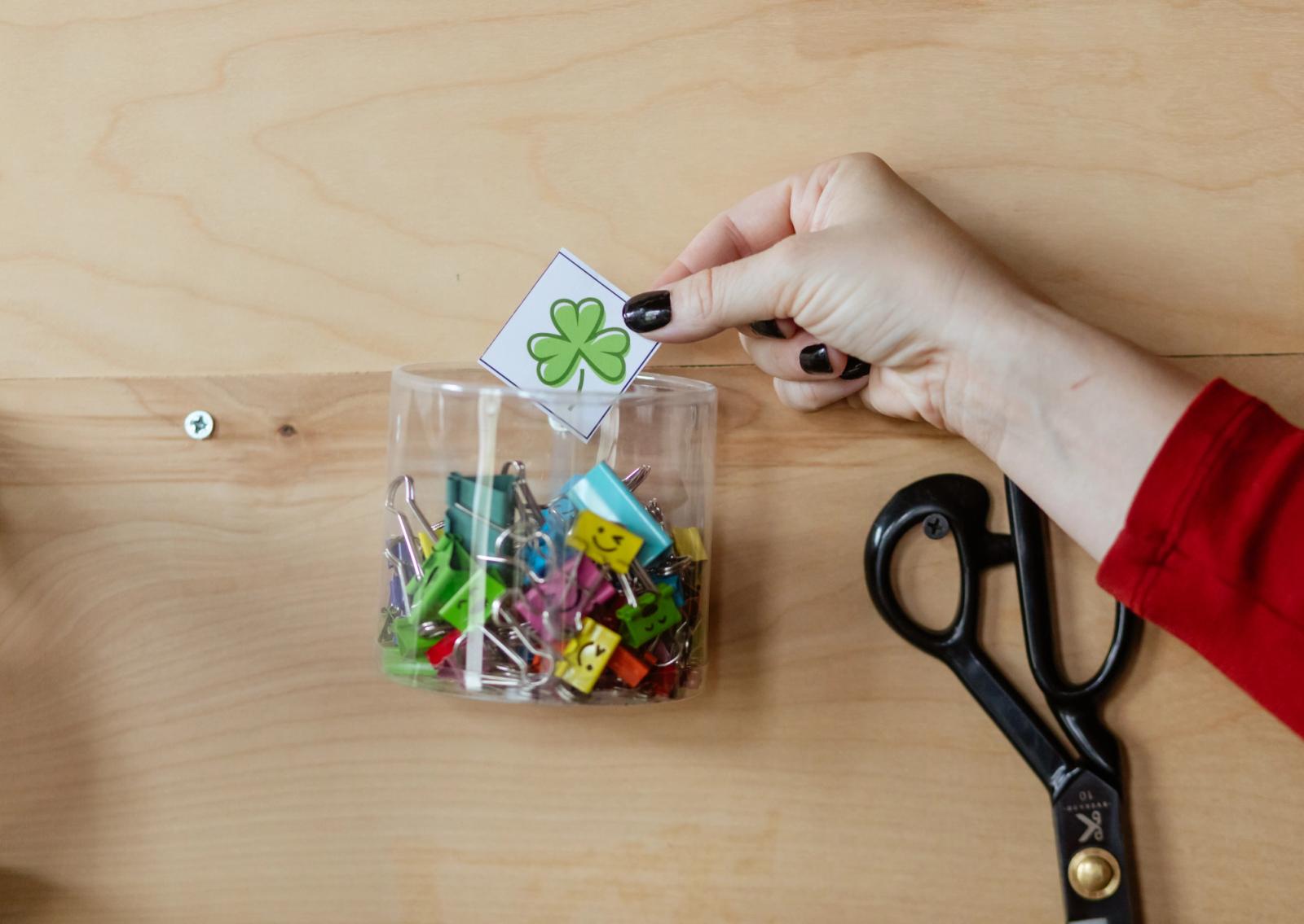 A hand places a small piece of paper with a green shamrock on it in a gar of paper clips.