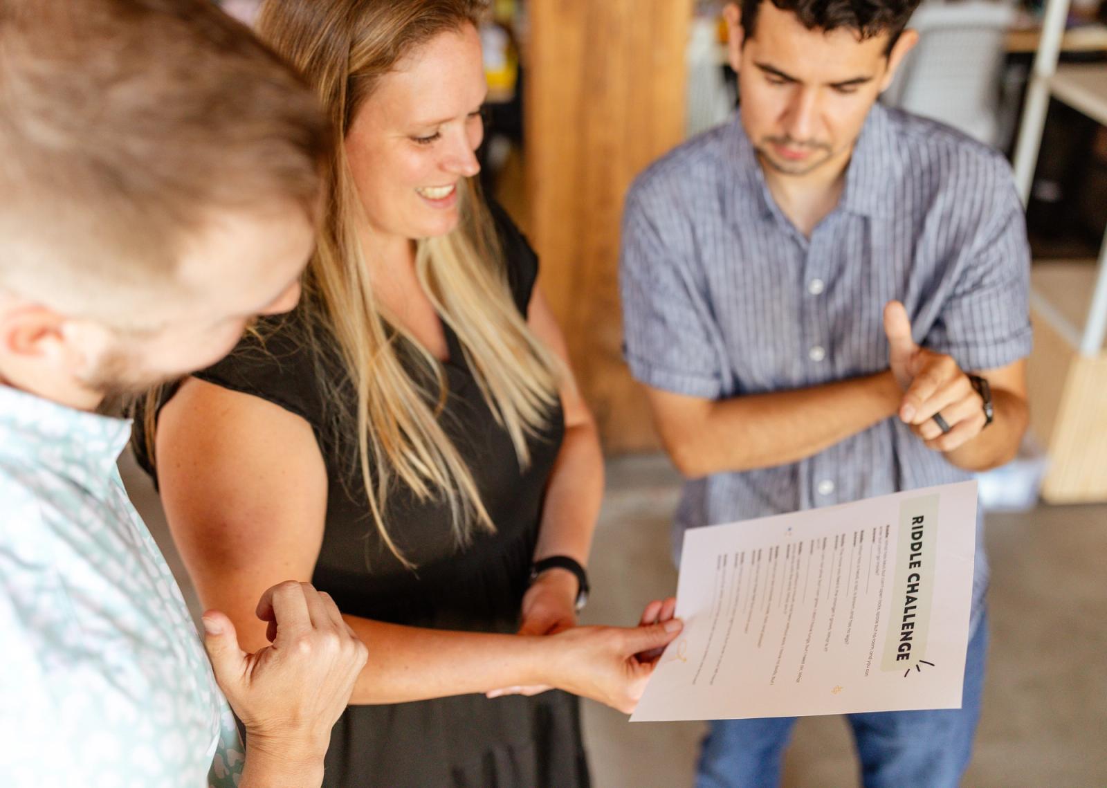 A woman holds a piece of paper. Two men stand on each side of her pointing at the paper.