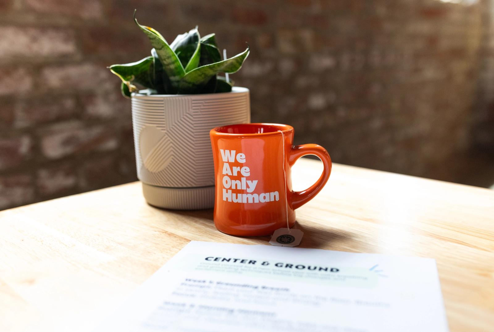 An orange coffee mug with a tea bag in it and a potted plant sit on a wooden table next to a sheet of paper. There is a brick wall in the background.