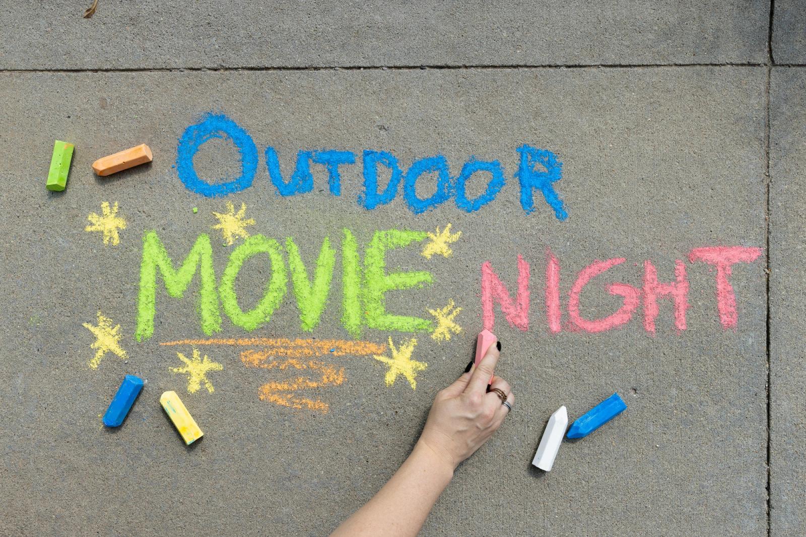 A person writes on the sidewalk with chalk. Message says "outdoor movie night."