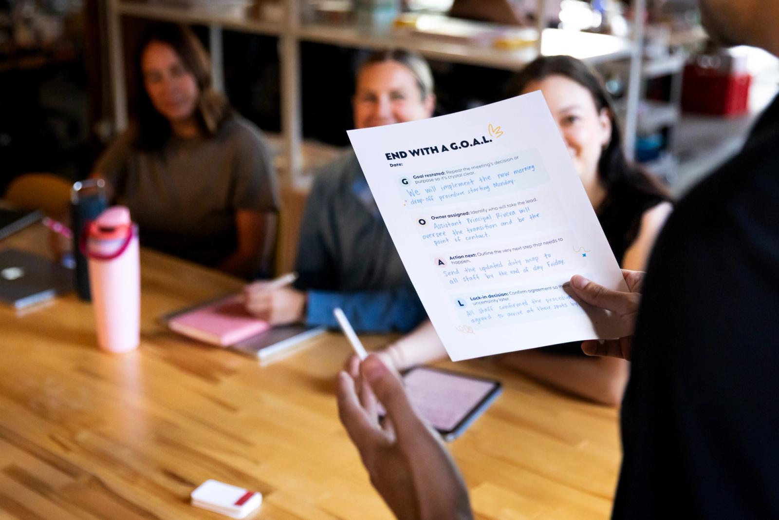 Man holds a piece of paper with writing on it as he stands in front of three women seated at a table.