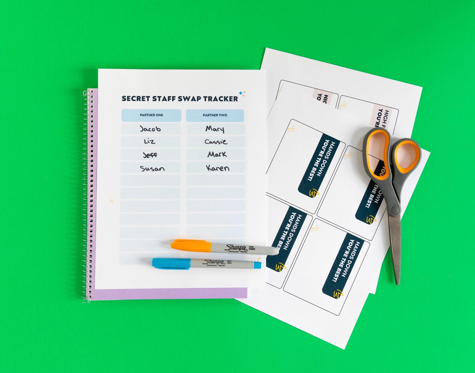 A purple notebook with a paper sign-up sheet and markers on it lay on a green background. Next to it are sheets of paper and a pair of scissors.