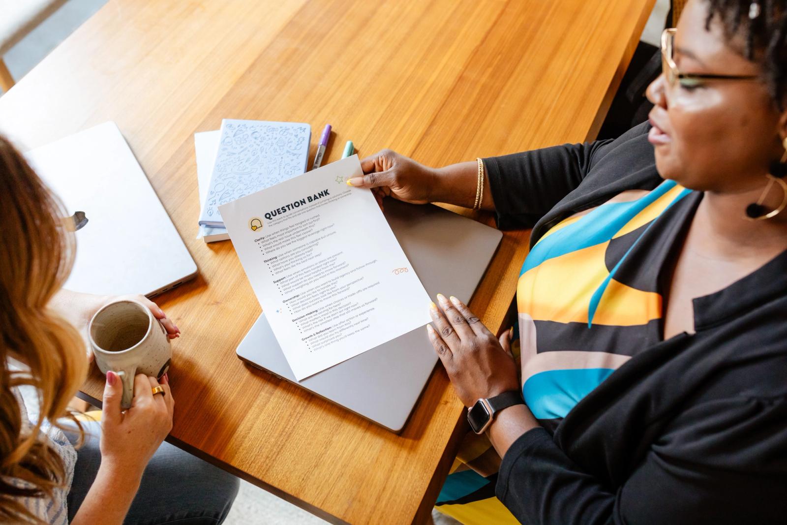 One woman reads a sheet of questions to another woman sitting at a table. 