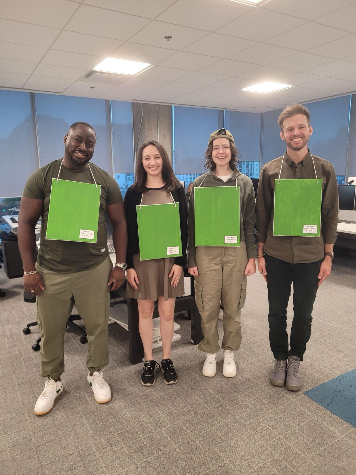 Four people in an office, each wearing a green board around their neck labeled “Submit compliance review,” dressed as Greenboard for Halloween.