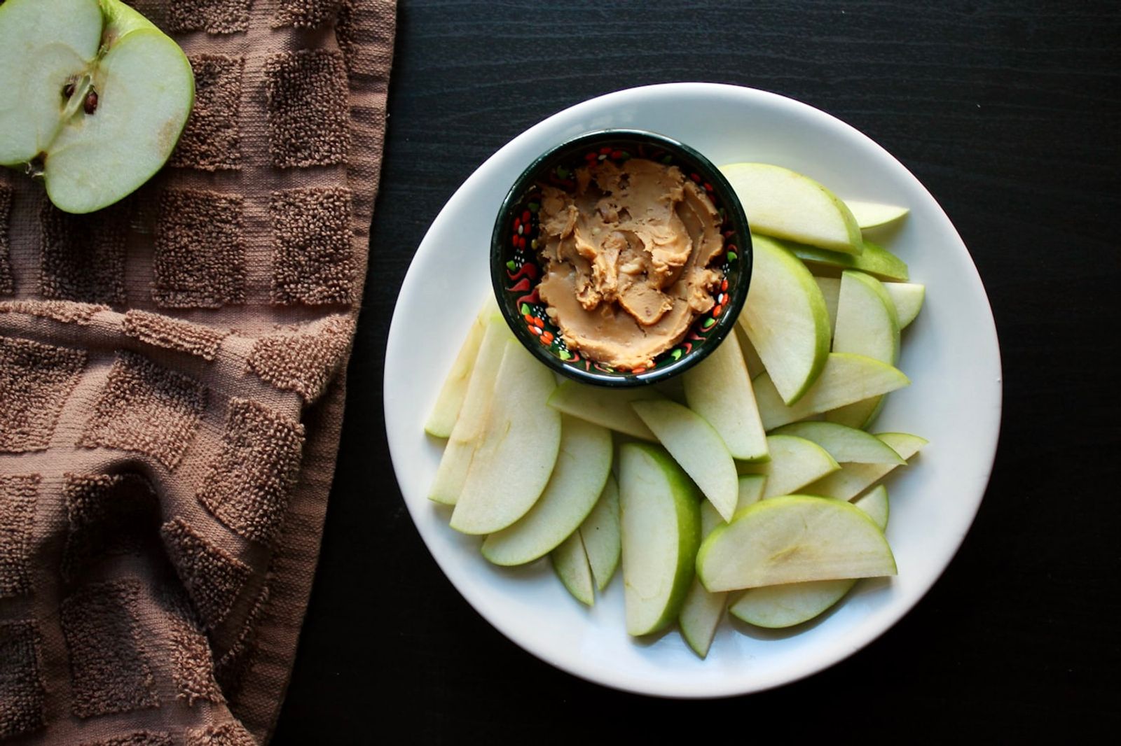 A sliced apple arranged with a small bowl of peanut butter on a wooden board, photo by Aasiya Khan on Unsplash