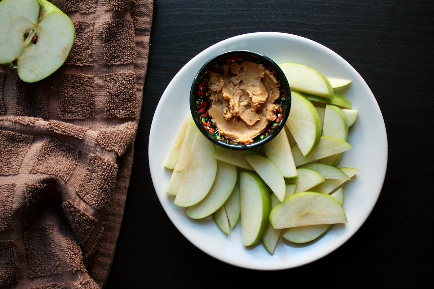 A sliced apple arranged with a small bowl of peanut butter on a wooden board, photo by Aasiya Khan on Unsplash