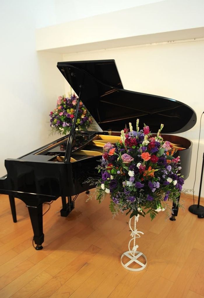 A grand piano with tall floral arrangements beside it in a white-walled hall