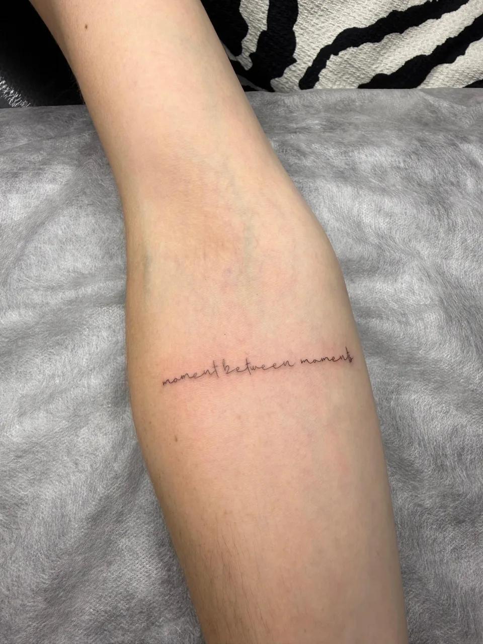 Elegant minimalist script tattoo reading 'moment between moments' in fine line cursive font, custom-inked at GG Tattoo studio in Budva, Montenegro.