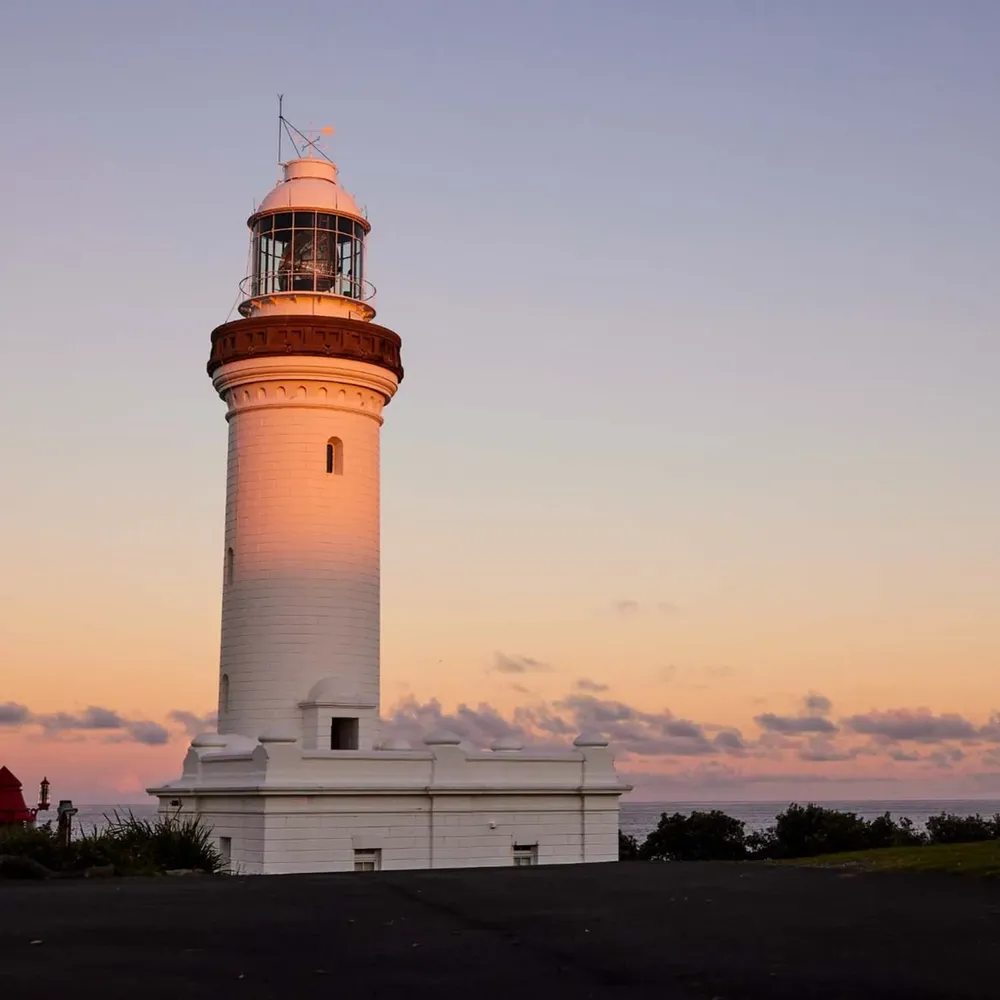 White lighthouse near Reflections Holiday Parks, standing tall at sunset with a pastel orange and purple sky in the background
