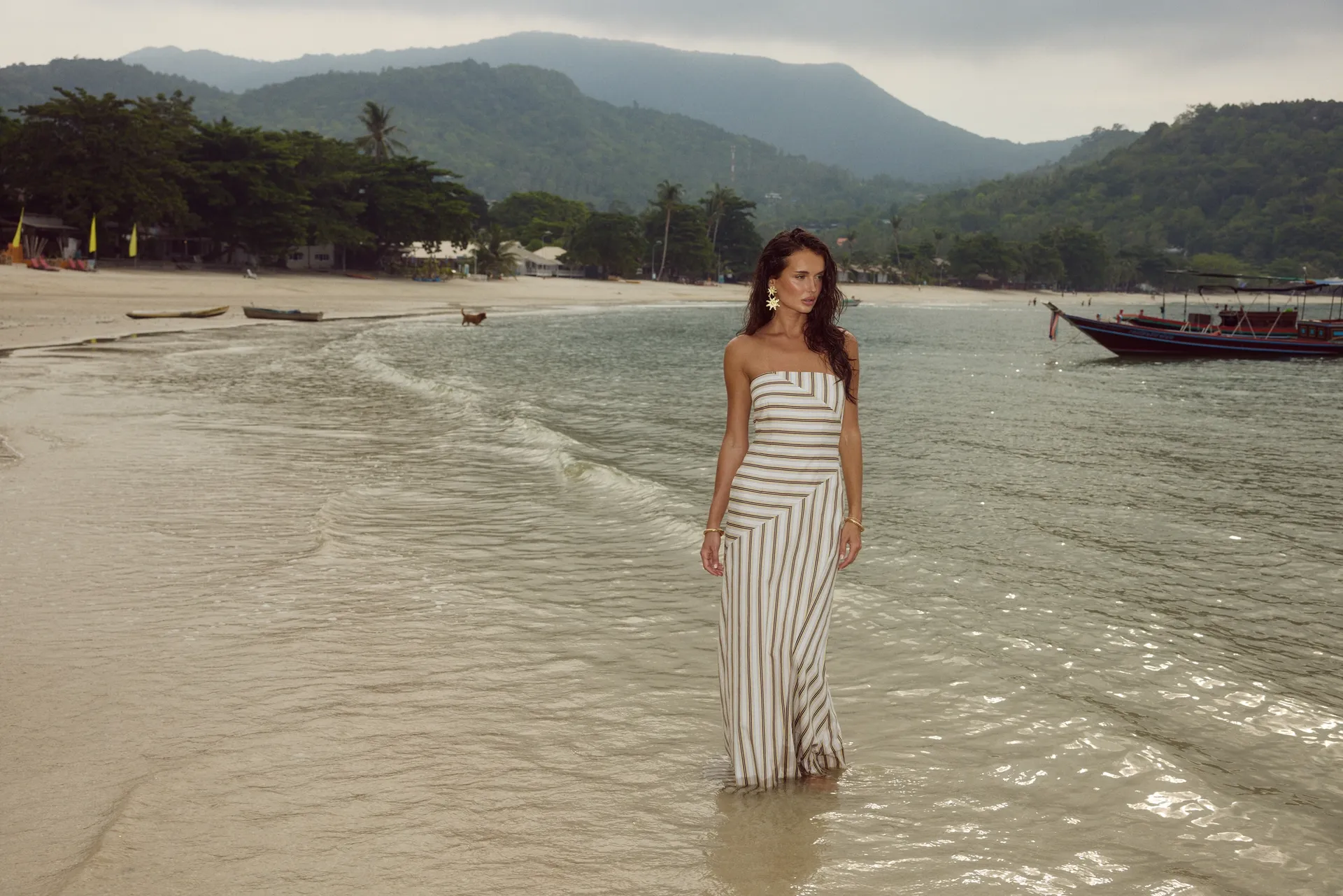 Model wearing Runaway The Label strapless striped maxi dress walking along tropical beach with ocean and mountains in background