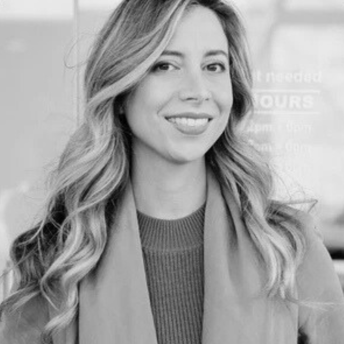 Roxy from Dress For A Night smiling in a black and white portrait, wearing a knit top and coat with loose wavy hair.