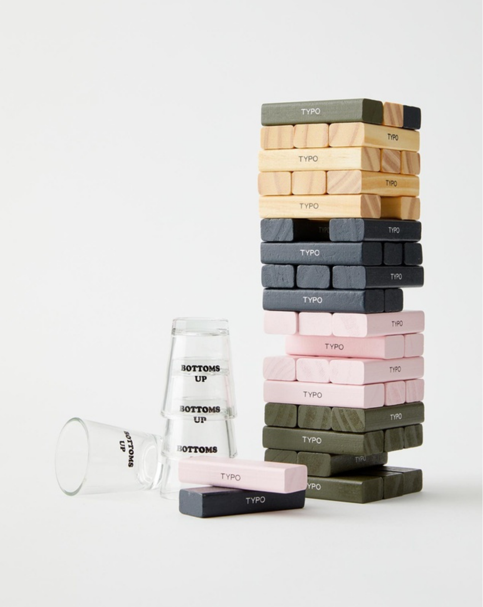 Typo drinking game set featuring colorful Jenga-style blocks in pink, grey, green, and wood tones, with “Bottoms Up” shot glasses on a white background
