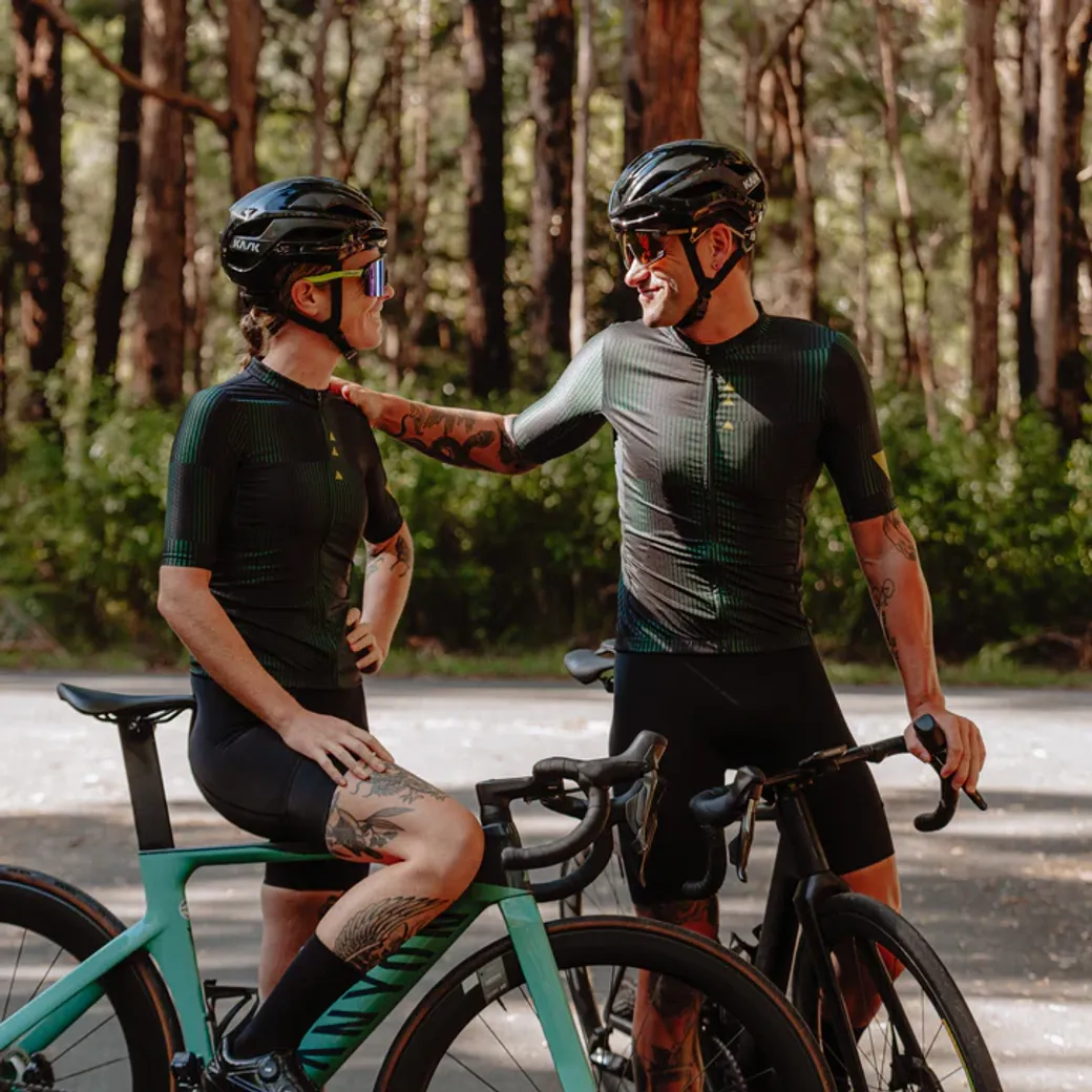 Two cyclists wearing matching black and green NeoPro cycling kits, helmets, and sunglasses, smiling and chatting while resting on their bikes in a forest road setting