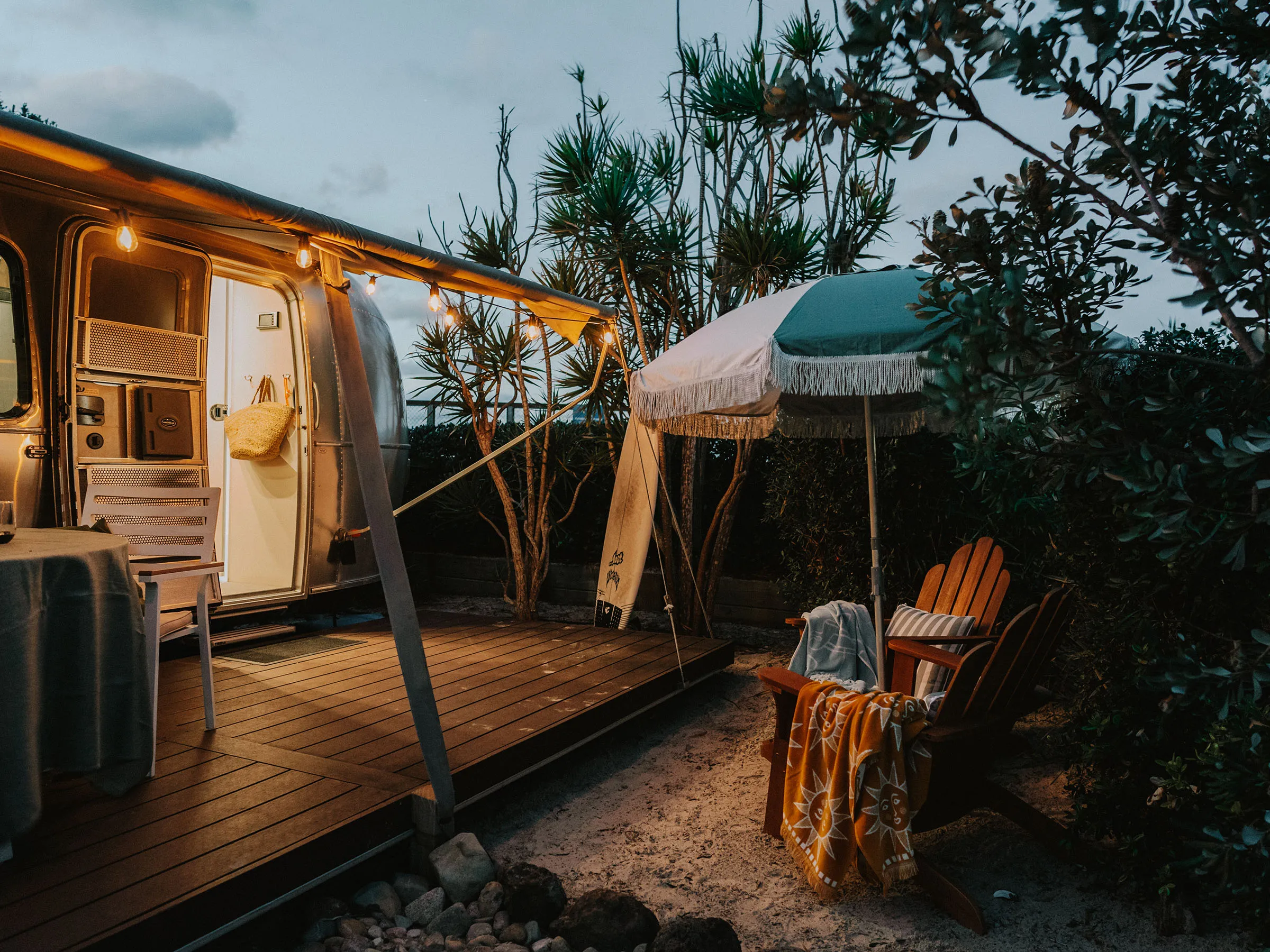 Reflections Holidays glamping site with a retro airstream, deck chairs and a fringed umbrella lit at dusk.