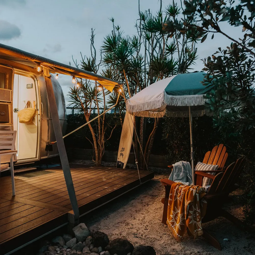 Reflections Holidays glamping site with a retro airstream, deck chairs and a fringed umbrella lit at dusk.
