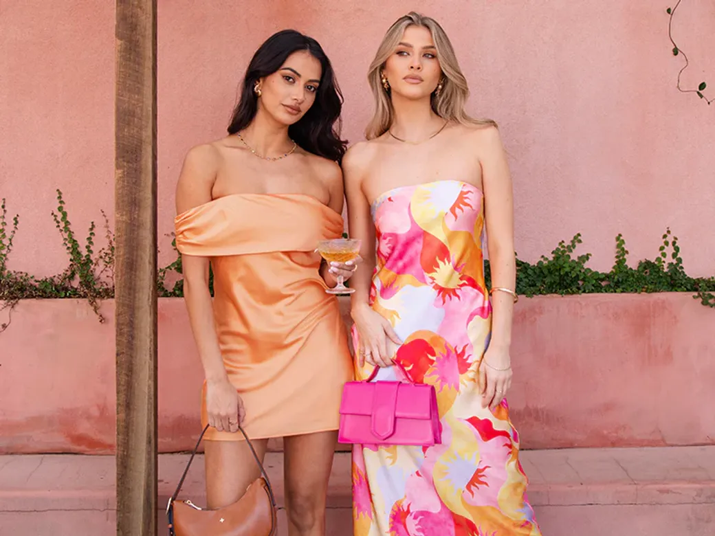 Two women posing against a pink wall, wearing vibrant Beginning Boutique dresses – one in a satin orange mini dress, the other in a strapless floral maxi dress holding a bold pink handbag