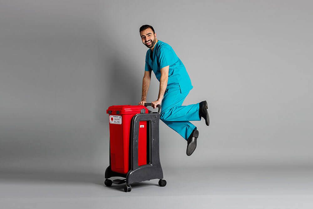 Hospital staff member in teal scrubs playfully riding a Daniels Health's red clinical waste bin on wheels in studio
